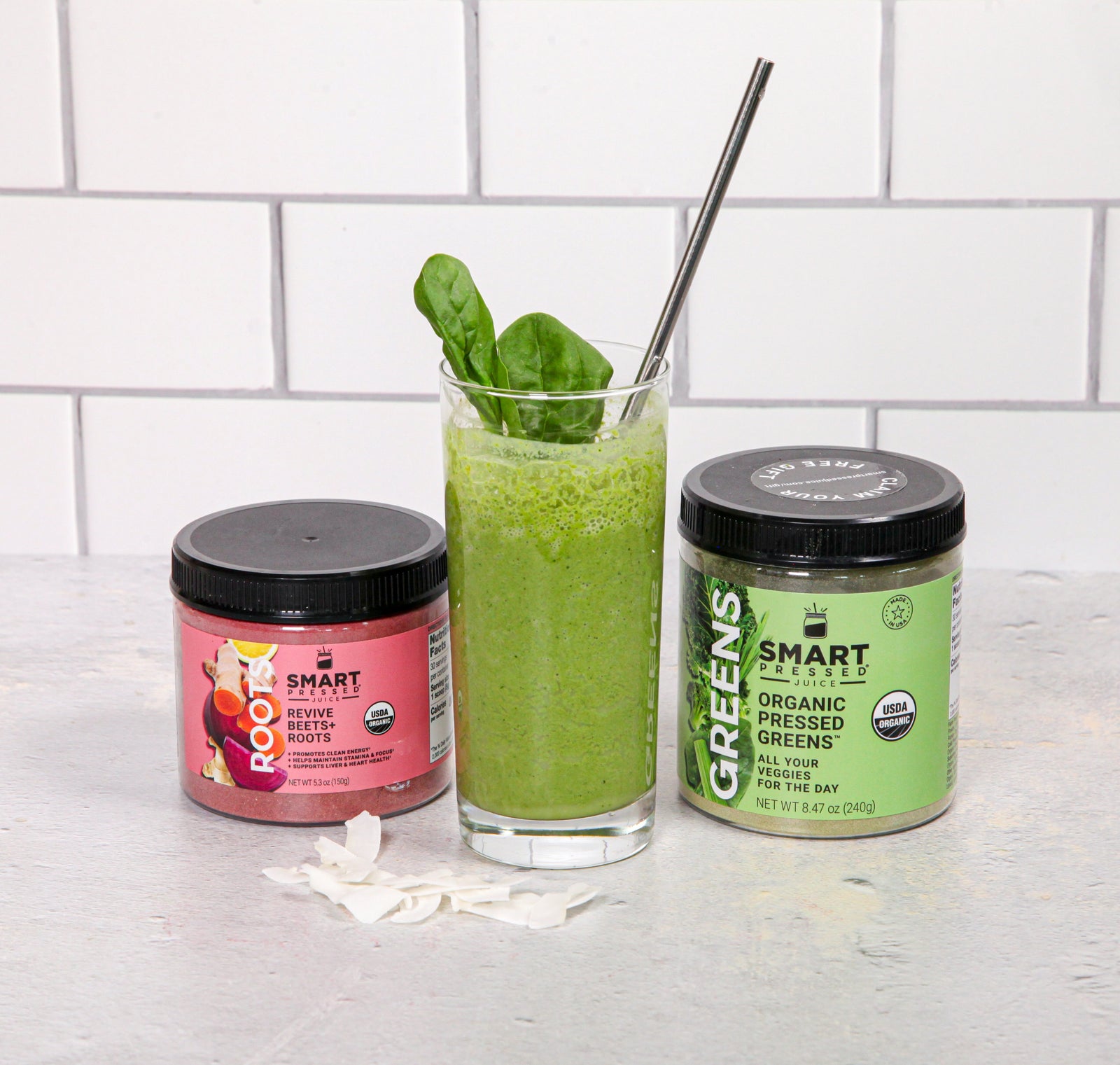 two smart labeled jar with a glass in the middle filled with cherry greens smoothie topped with two spinach and a black patterned plastic straw surrounded by coconut chips on top of a white table