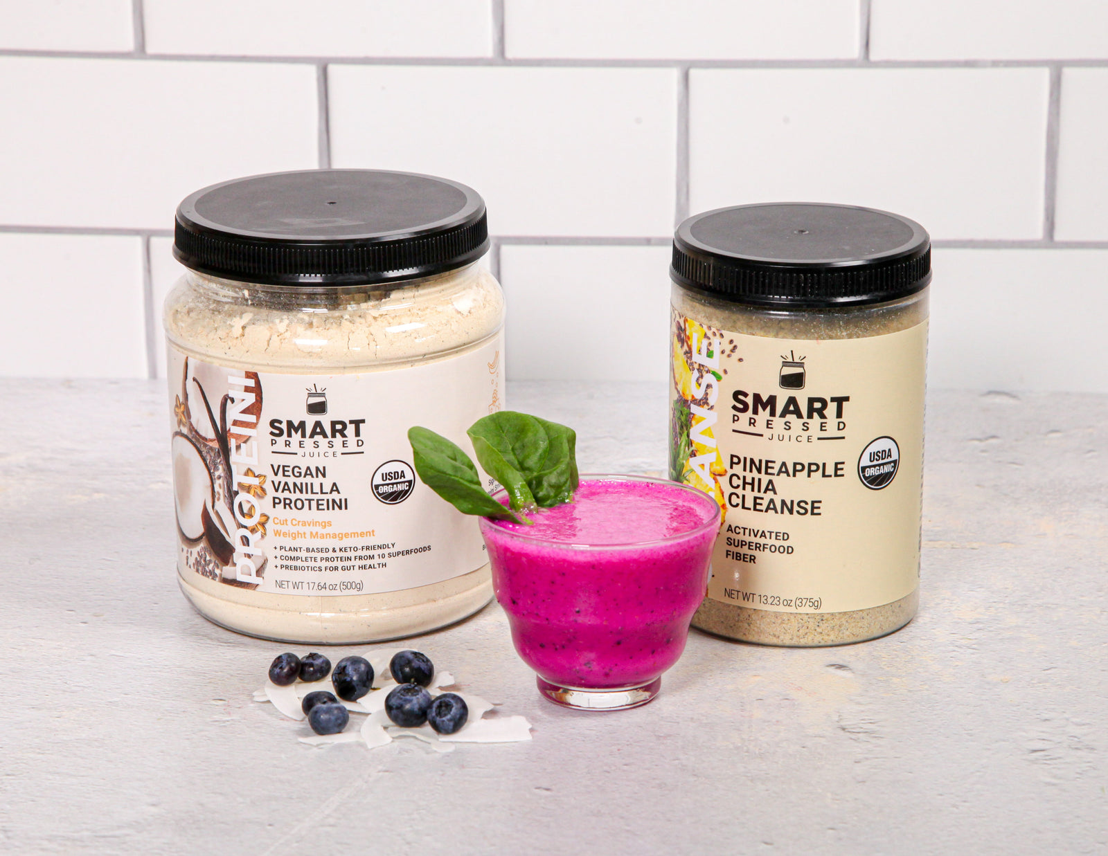 1 jar of Vegan Vanilla Proteini beside a glass of Amy's Dragonfruit Smoothie garnished with green leaf beside a jar of Pineapple Chia Cleanse and on the surface are blueberries. Set against a brick wall.