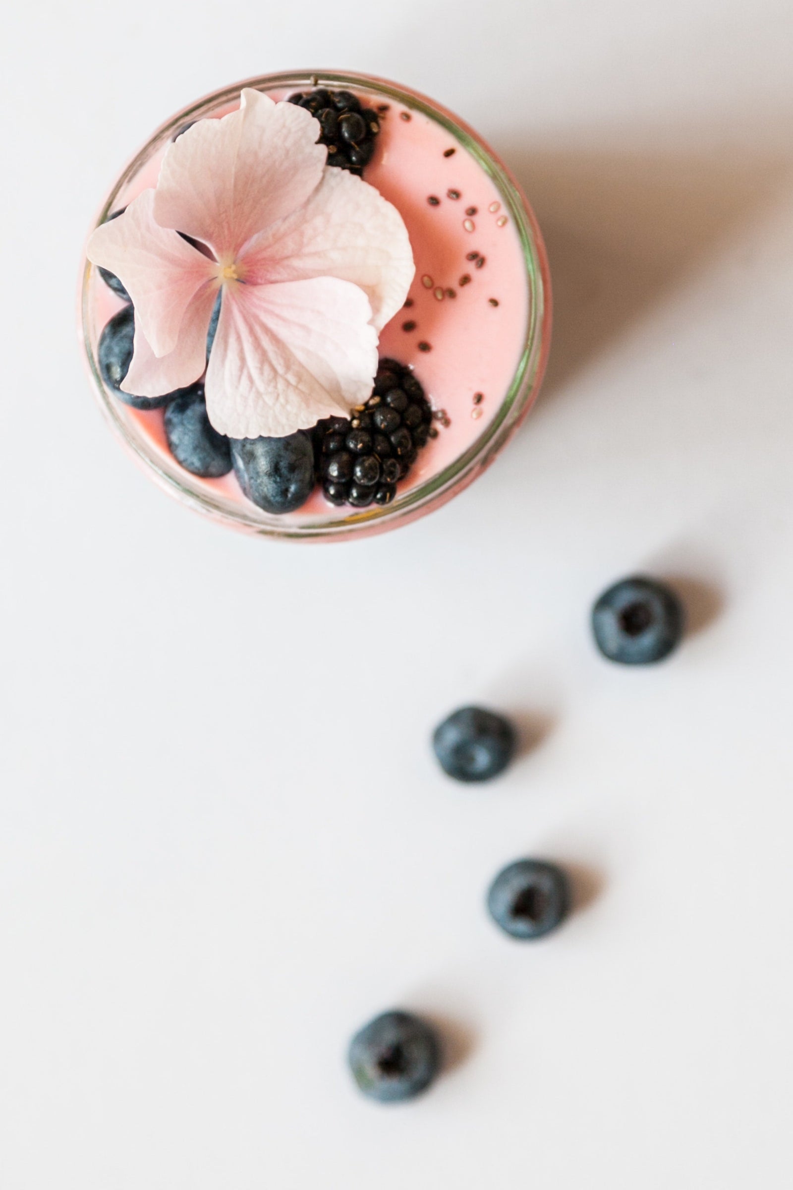 top view of a bowl of wildberry pineapple chia cleanse topped with berries and roasted sesame seeds and a pink flower on top with four berries on the side on top of a white table