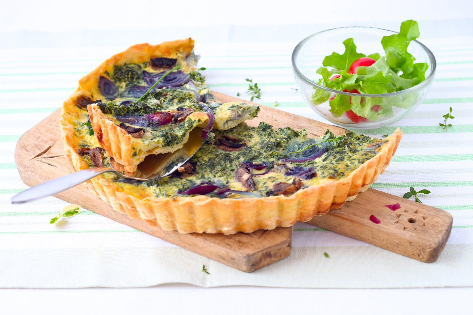 Asparagus Quiche with Tarragon Vegan pie on a wooden cutting board with a spoon and pie on top, beside is a small bowl of lettuce salad with cheery tomatoes.