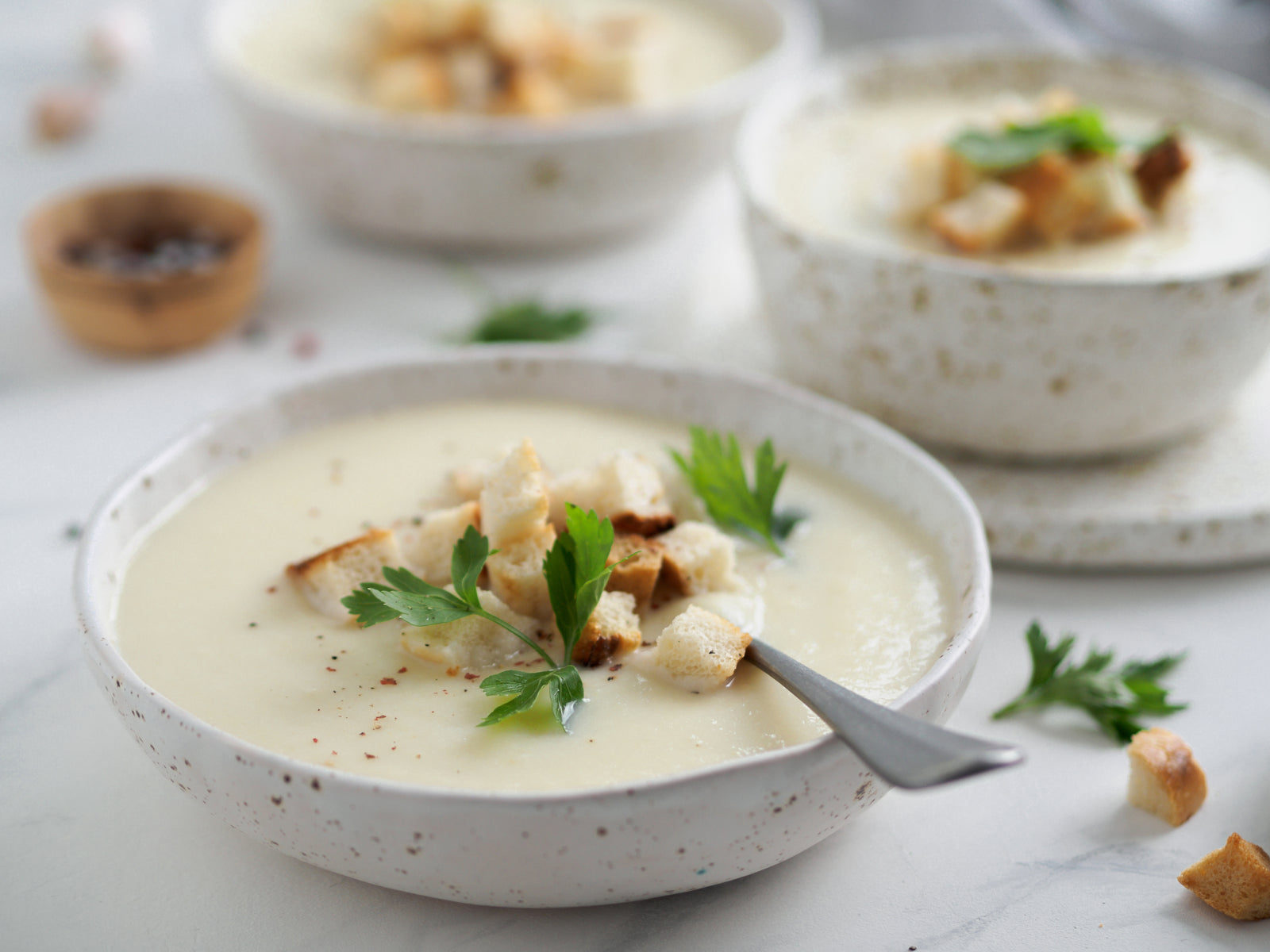 side view of three bowls of creamy cauliflower & white bean soup with garlic croutons topped with croutons and parsley on top of a white table