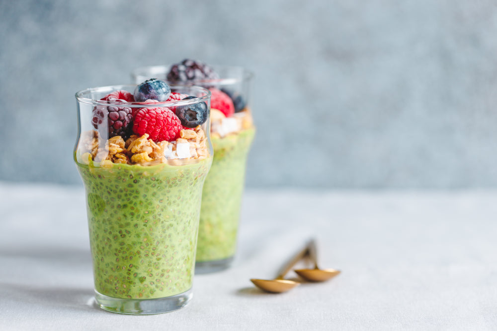 two glasses of Chia Pudding with Matcha Whip topped with berries and nuts with spoon on the side on top of a light blue table