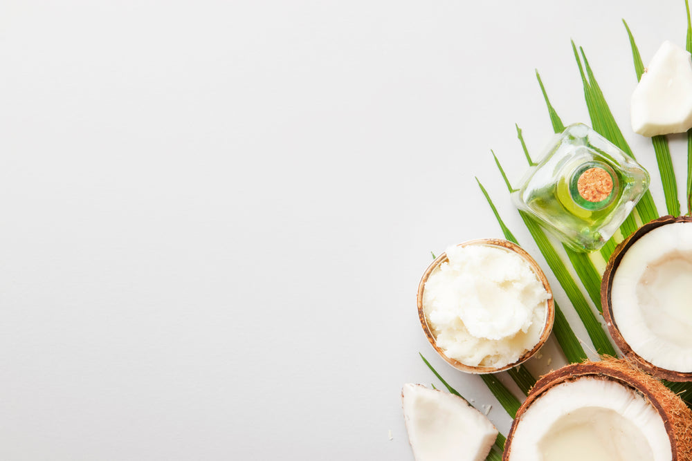 top view of a cut of coconuts and a bowl of scraped coconut meat and a bottle of coconut oil and slices of coconut meat