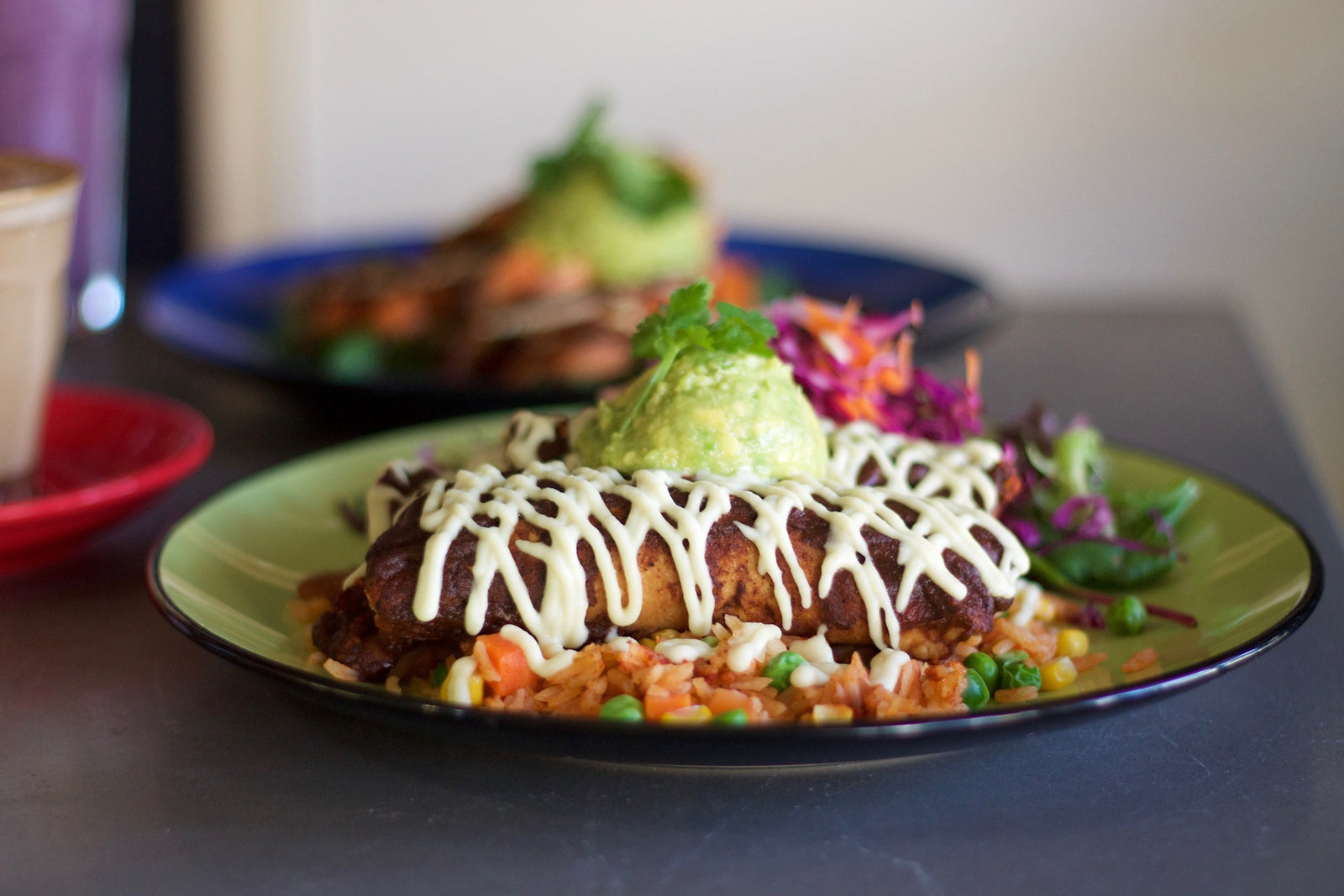 a plate of potato and kale tortilla in an enchilada sauce drizzled with mayonnaise and topped with a scoop of guacamole with a green salad side dish. Behind is another plate of the same content.