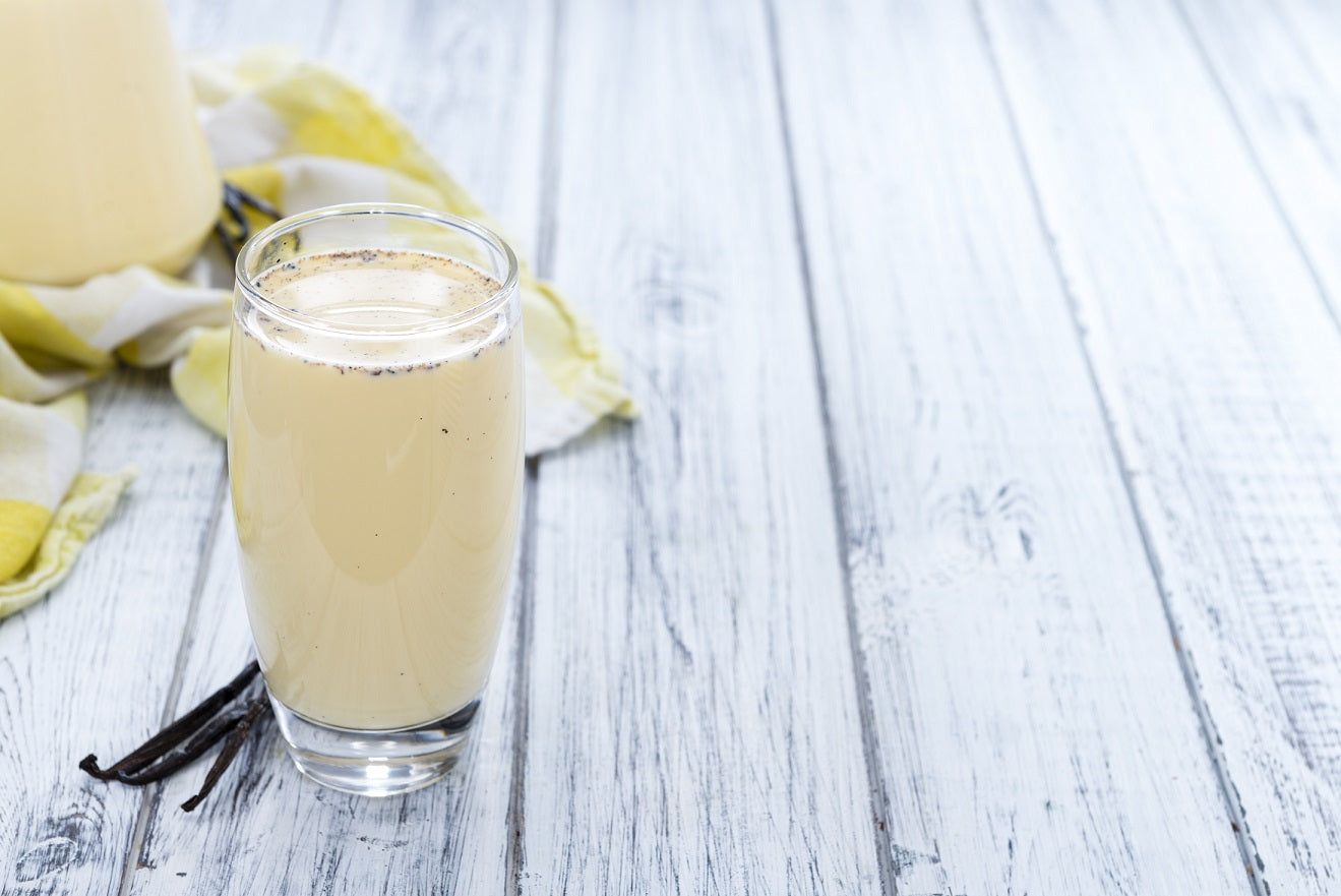 1 glass of Vegan Vanilla Proteini beside a stick of vanilla, behind is a yellow and white cloth on a wooden surface.