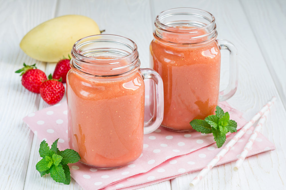 two jars of vanilla strawberry mango smoothie on top of polka dot pink white table clothe surrounded by mint leaves, two pink white paper straw, three strawberries, and one mango