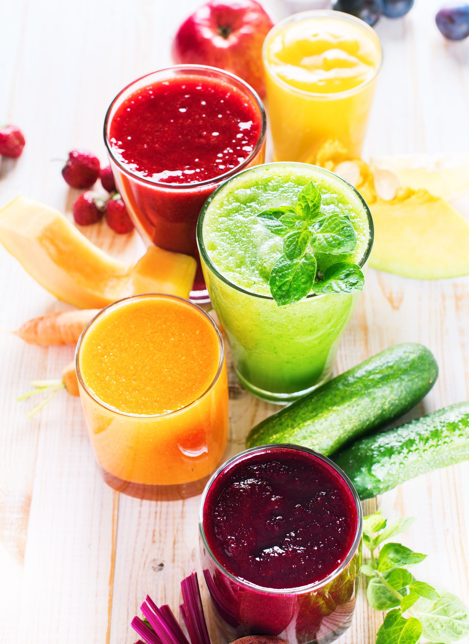 five glasses of different smoothie surrounded by apple, grapes, berries, carrots, cantaloupe, and two pickles