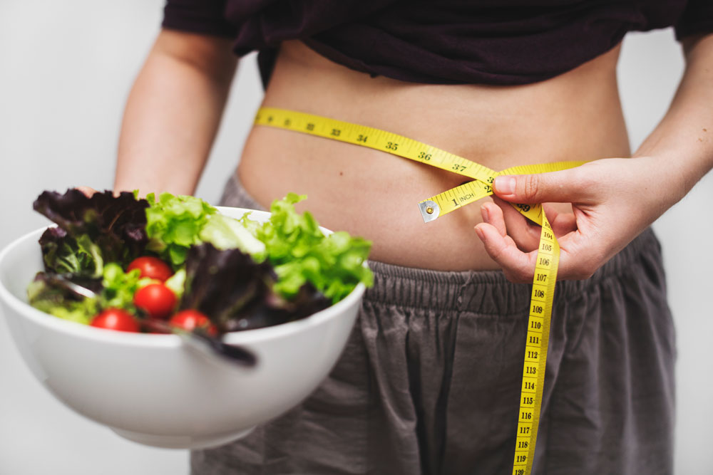 Image of a half-body woman holding a bowl of salad on her right hand and a measuring tape on her waist using her left hand.