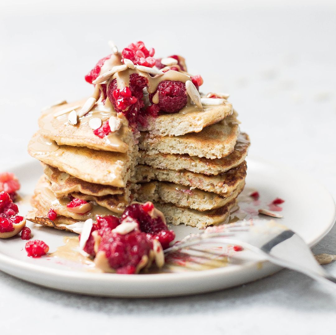 six-layered banana proteini pancakes with sliced portion topped with berries and squash seeds on top a plate surrounded by berries, squash seeds, and fork on the side