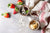 top view of vanilla proteini mug cakes, one mug with a spoon with a red patterned table cloth, and four strawberries and white chocolate chips on the side on top of a marble table