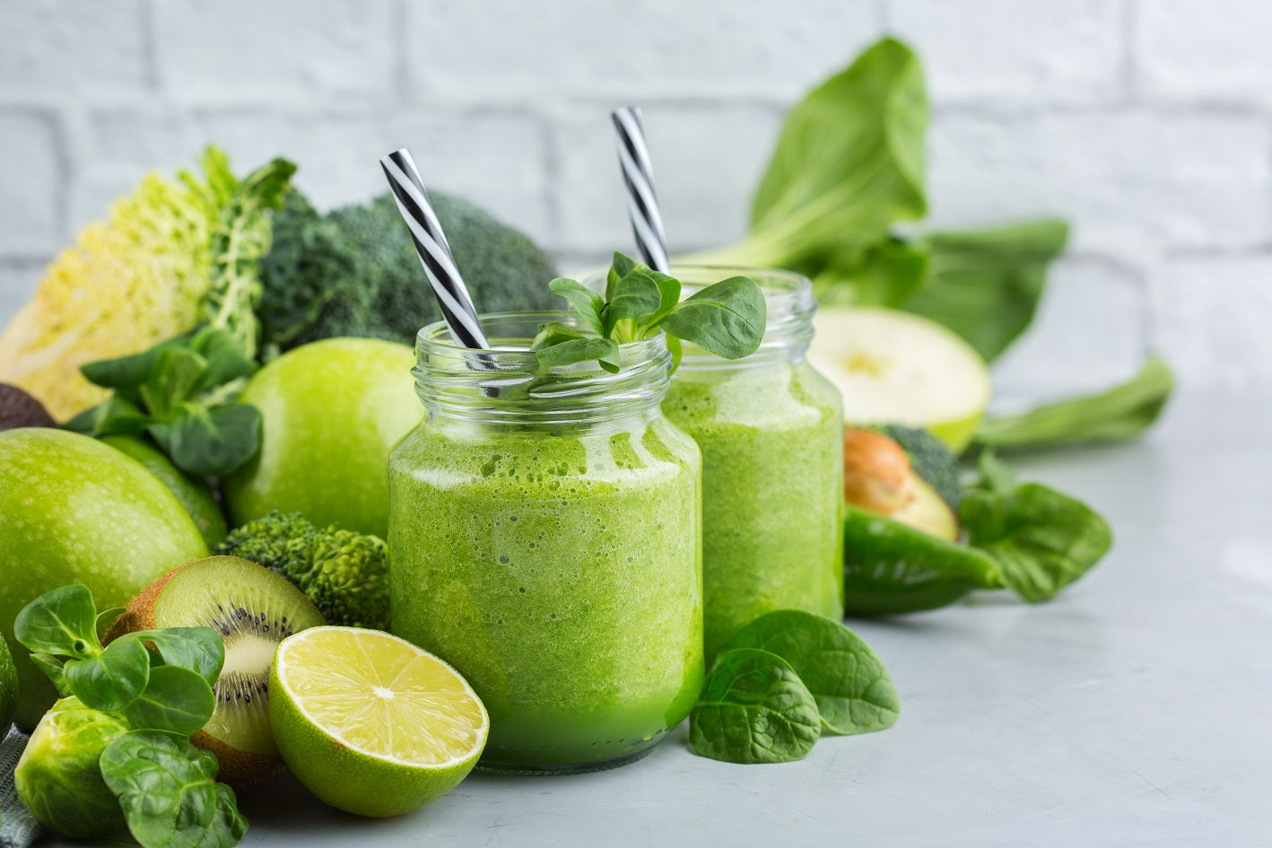 two jars of organic pressed greens with two black patterned staw topped with arugula surrounded by a sliced of lime, sliced kiwi, broccoli, three green apples, arugula and bokchoy