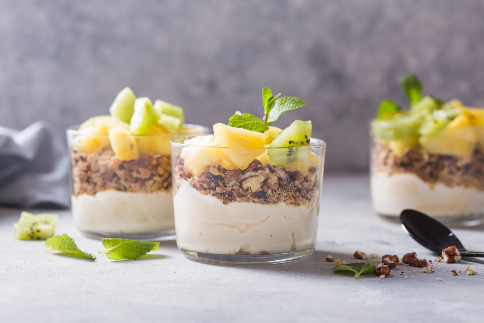 three glasses of pina colada overnight oats topped with nuts and slices of pineapple and kiwi with mint surrounded by mint, nuts and slices of kiwi on top of a table with powder blue table cloth