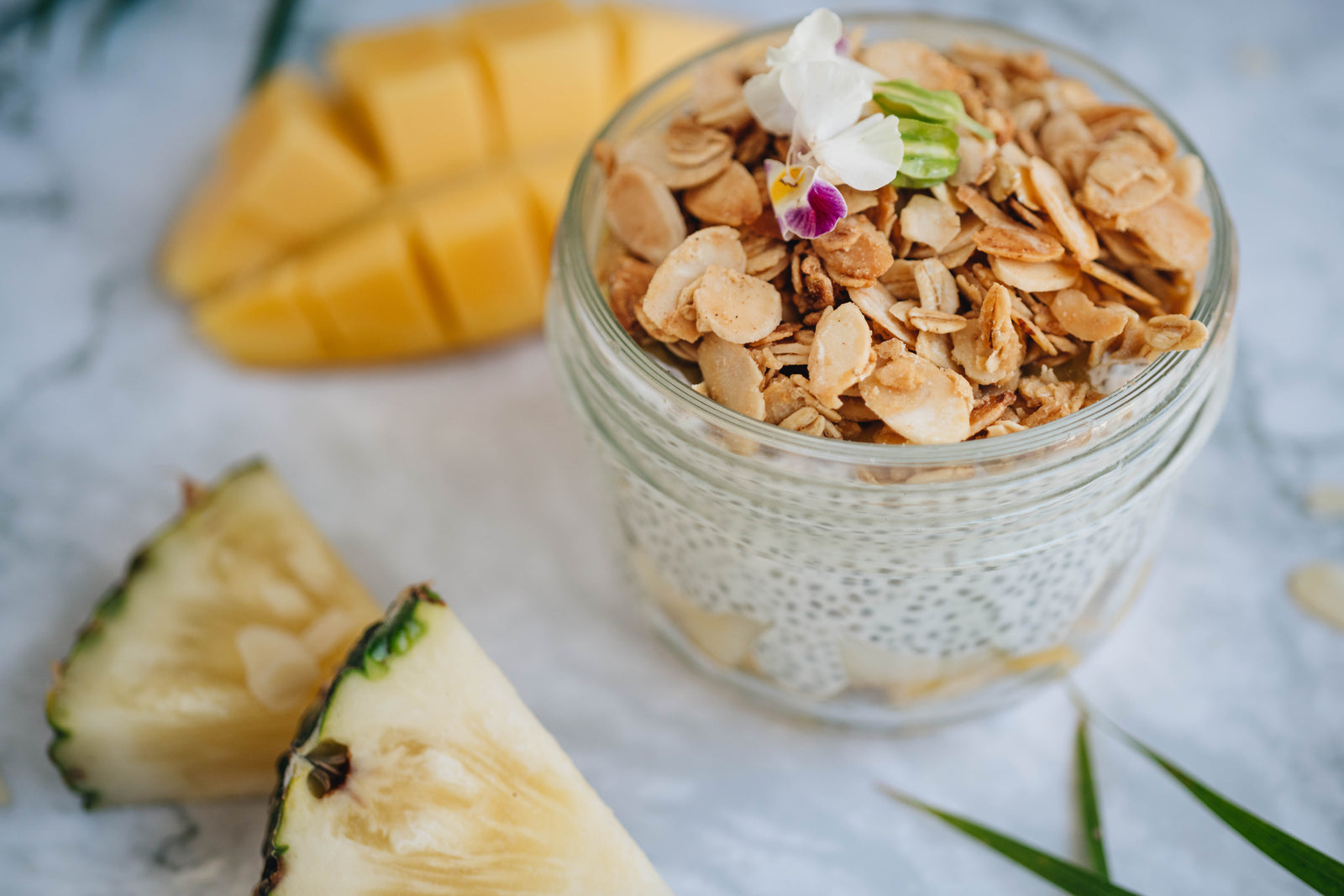 top view of a pineapple chia pudding jar topped with roasted nuts and flower with two slices of pineapple and a slice of mango cut into cubes on top of a marble table