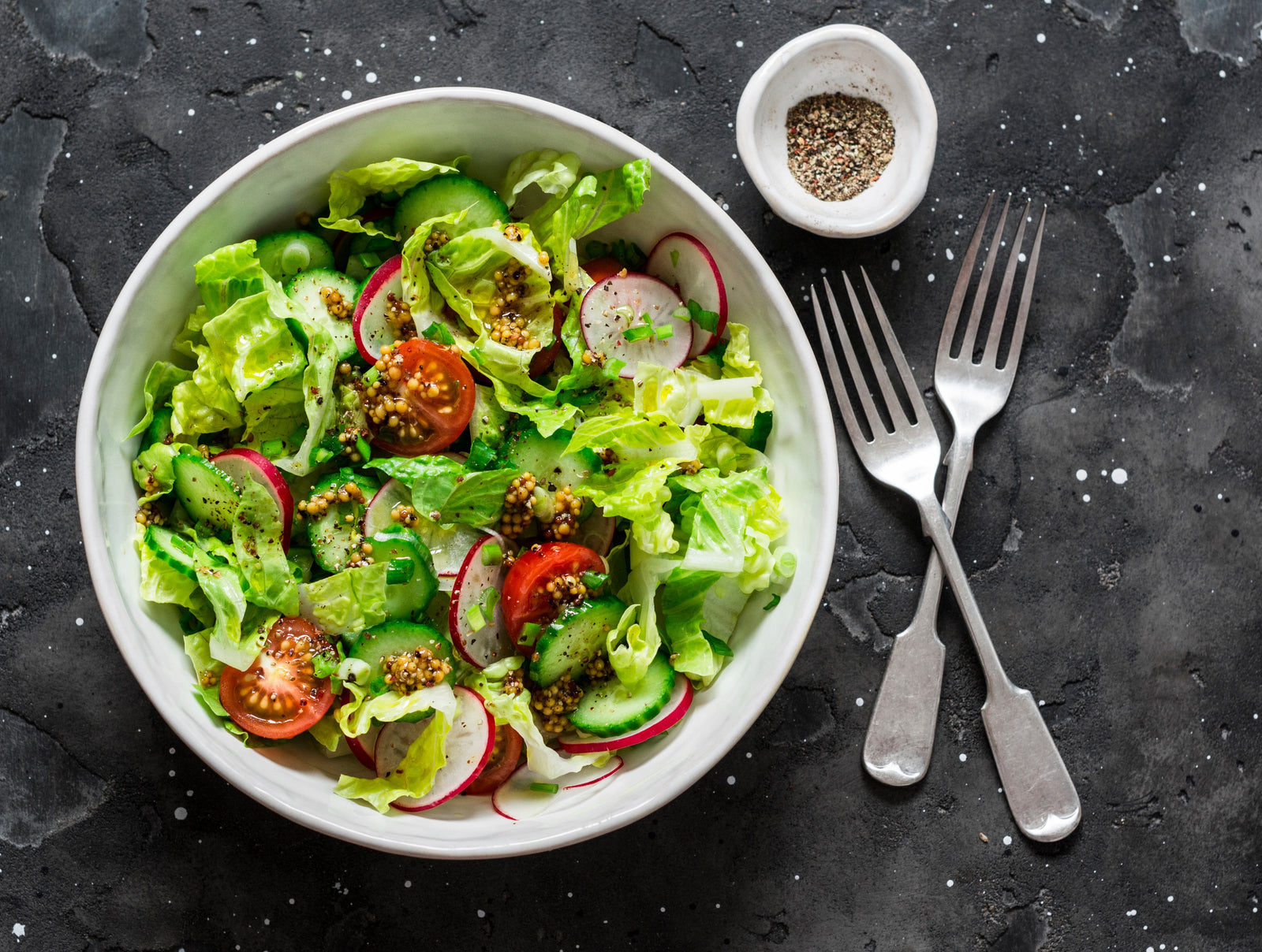 top view of a bowl with light Mediterranean salad with two forks on the side and a small bowl of crushed peppercorns on top of a black-painted concrete table