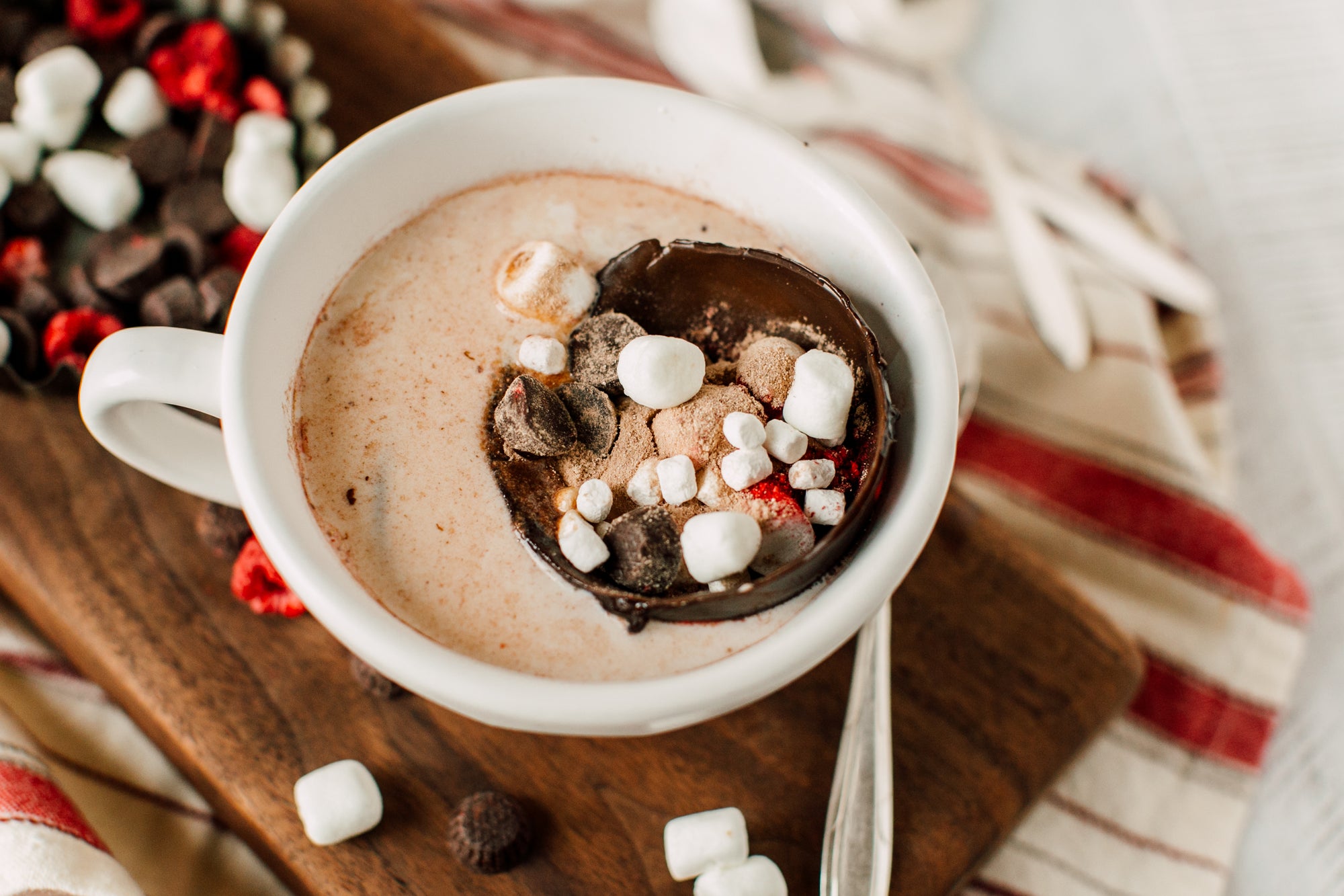 top view of a cup of melissa colwill's coco bombs topped with half-sphered chocolate with mallows, and chopped nuts inside with a spoon on the side and a paper molder full of chocolate and mallows on top of a wooden chopping board