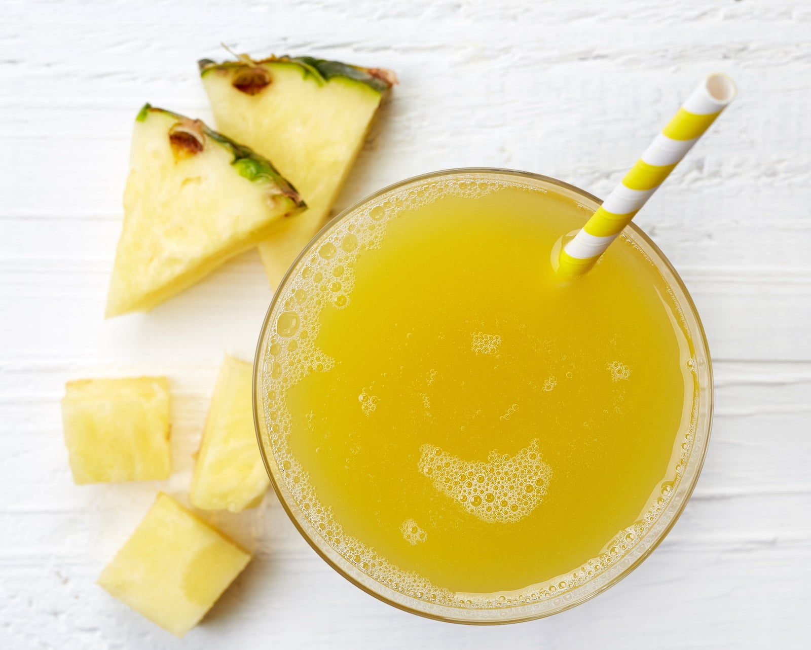 top view of a glass of sour pineapple chia with yellow patterned paper straw with slices of pineapple on the side on top of a white-painted table 