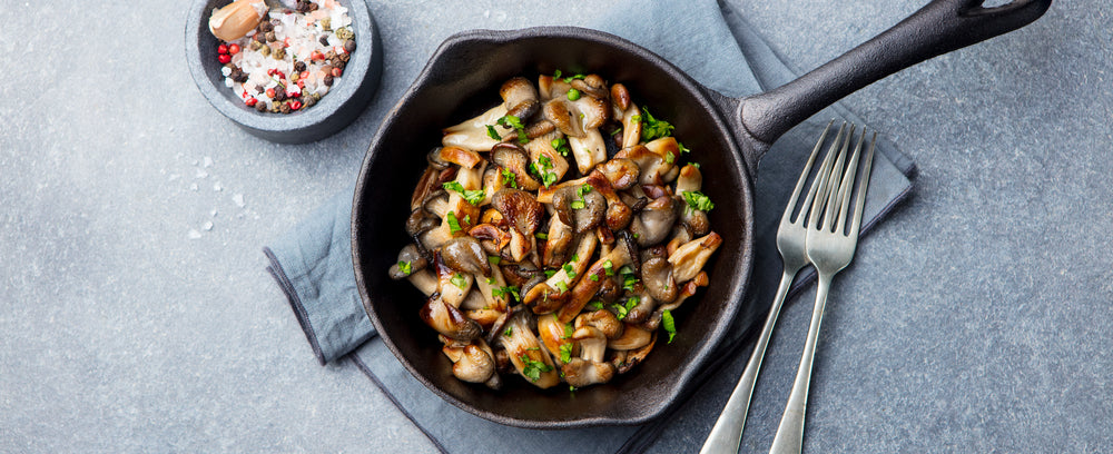 top view of skillet with cauliflower mushroom roast on top of a blue folded table cloth ith two forks on the side and a small bowl of mixed condiments on top of a blue-painted concrete table 