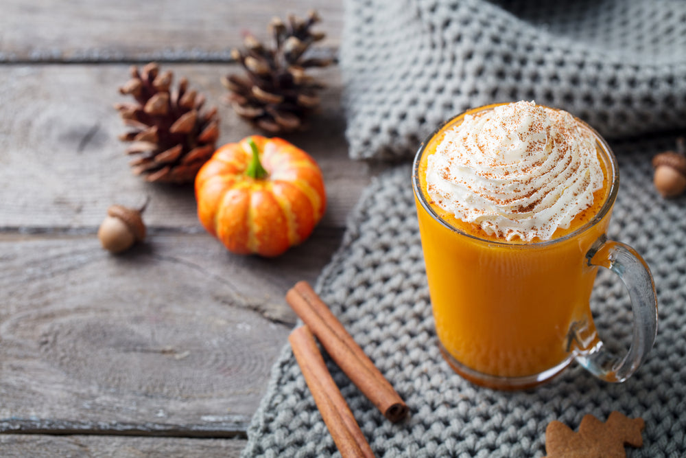 two glasses of pumpkin spice proteini with two small pumpkin and two spoons on the side with three cinnamon sticks on top of a weaved cloth  