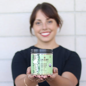 Zoomed-out woman holding a jar of 240 grams Organic Pressed Greens. She is standing against a brick wall.